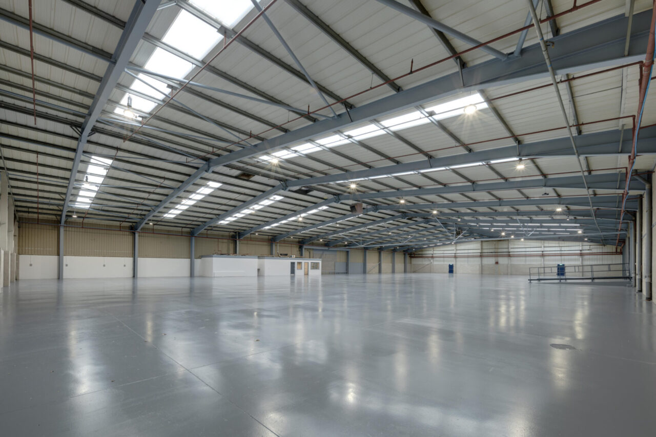 Large empty warehouse with polished floors, high ceilings, skylights, metal beams, and white partitioned rooms along one wall.