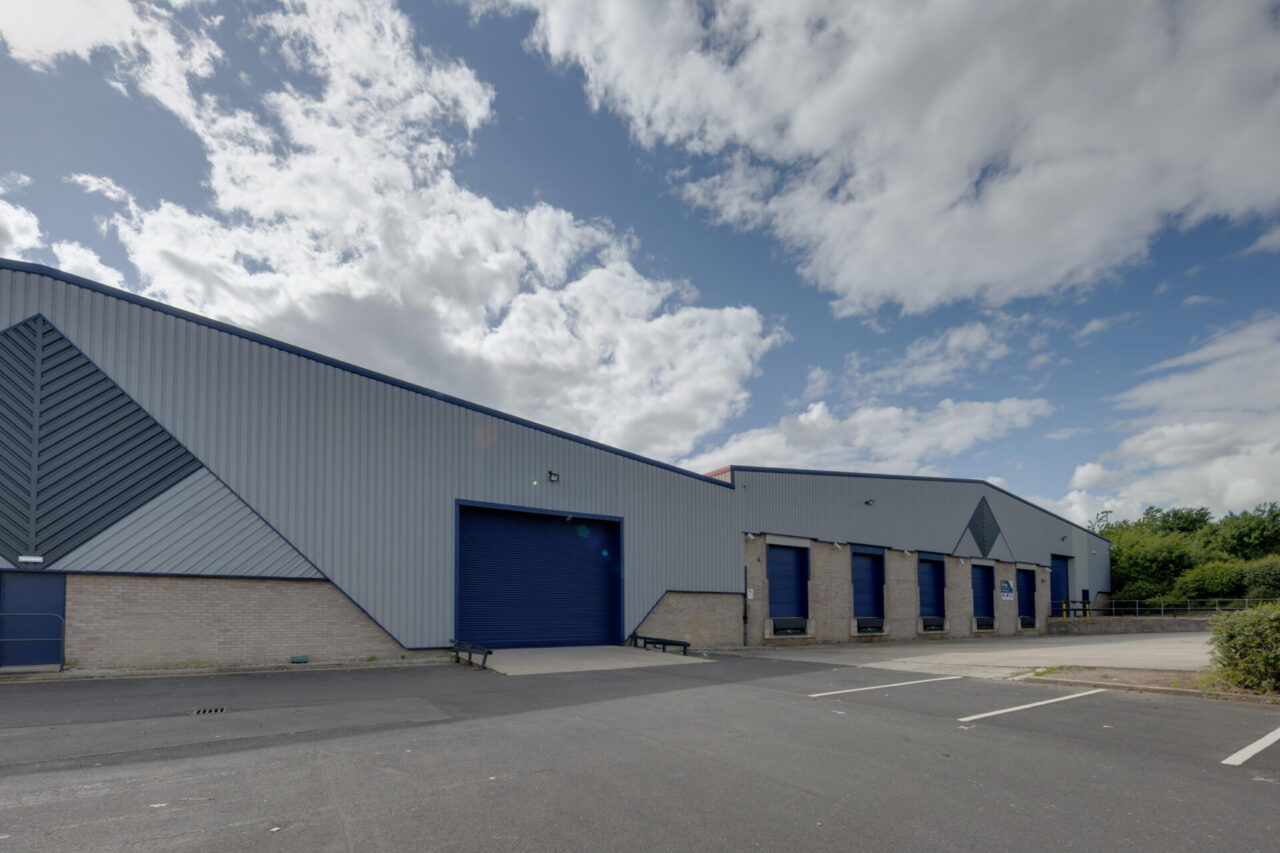 A large industrial warehouse with blue roller shutters, brick base, and metal cladding under a partly cloudy sky; empty car park in front.