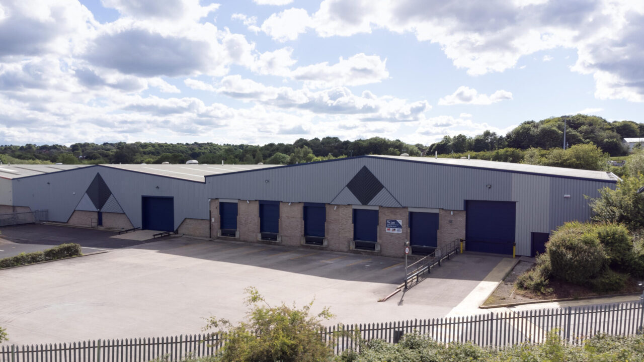 A large industrial warehouse with blue loading bays and a fenced yard, surrounded by greenery beneath a partly cloudy sky.