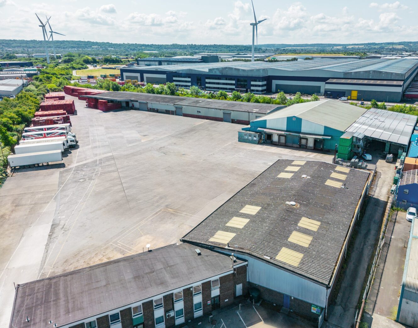 Aerial view of an industrial site showing warehouses, parked lorries, open concrete yard, and wind turbines in the distance.