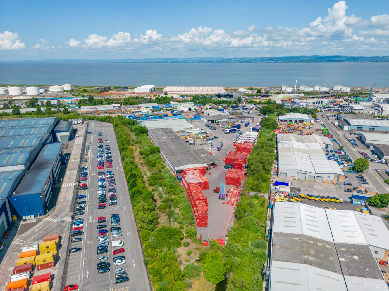 Aerial view of an industrial area with warehouses, parked cars, shipping containers, and a body of water under a partly cloudy sky.