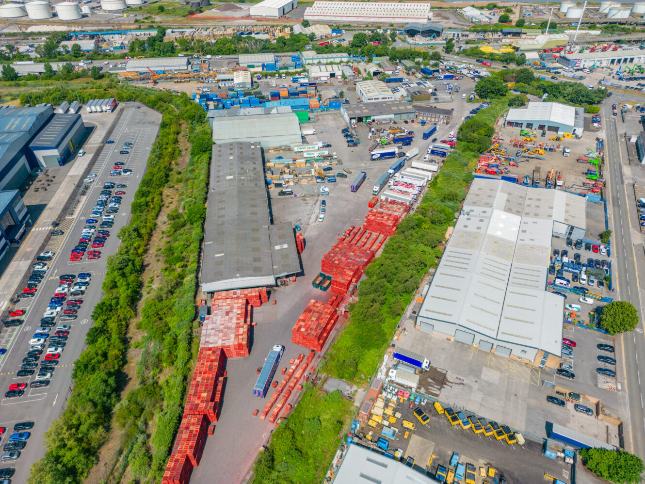 Aerial view of an industrial area featuring warehouses, parked vehicles, storage containers, and greenery in the surroundings.
