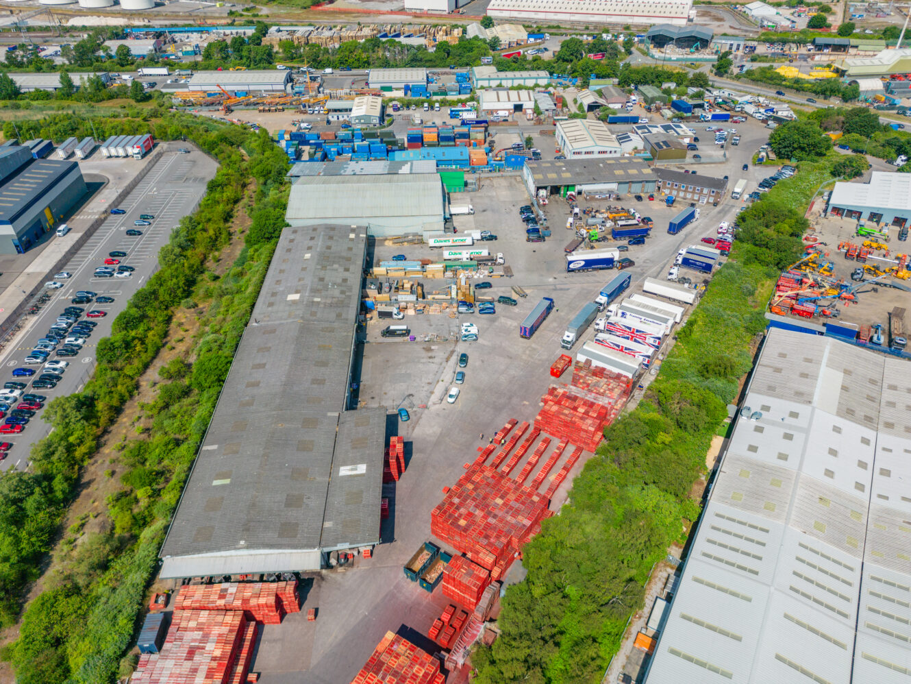 Aerial view of an industrial storage yard with warehouses, lorries, shipping containers, and red materials amid greenery and roads.