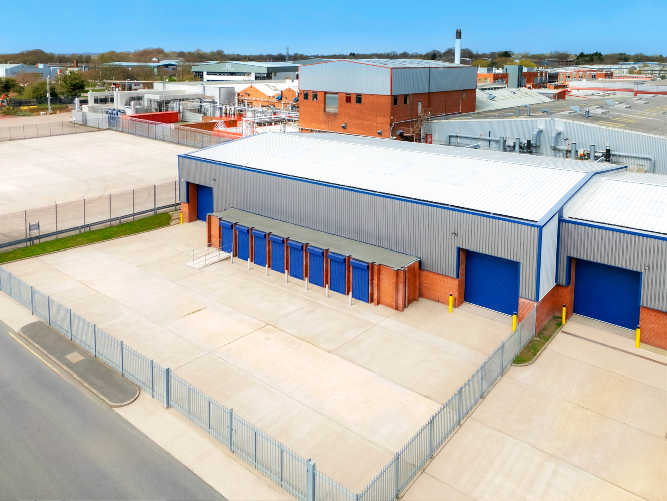 A fenced industrial facility with a large warehouse, several blue roller shutters, and an empty concrete yard. Other warehouses at the back.