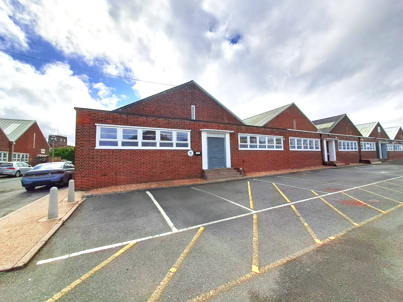 Single-storey red brick building with a sloped roof, white-trimmed windows, and blue door, beside marked parking under a cloudy sky.