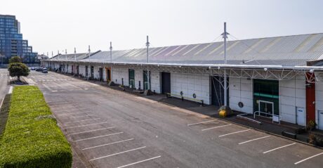 A row of industrial warehouse units with roller doors, vacant parking spaces in front, and a modern building in the background.