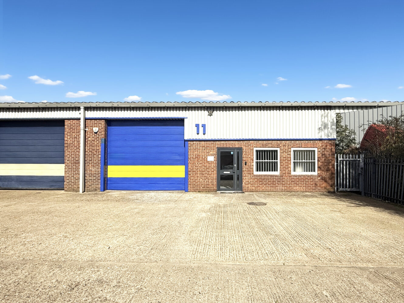 Single-storey industrial building with brick and metal exterior, blue and yellow roller shutter door, large garage door, entry door, and windows marked with the number 11.