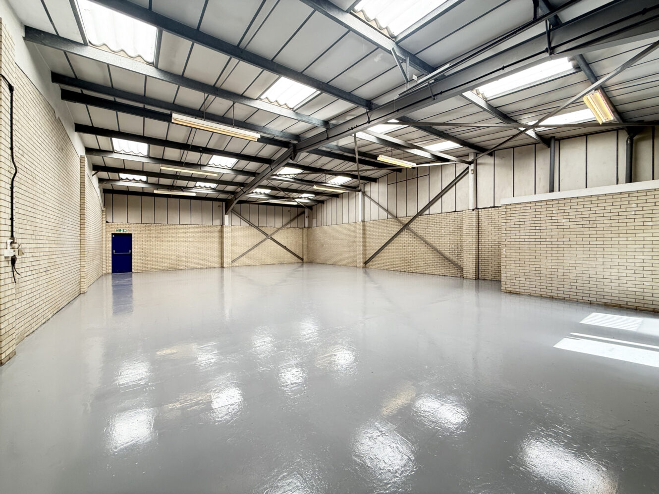Empty industrial warehouse with a shiny grey floor, exposed steel beams, cream brick walls, skylights, and a closed blue door.