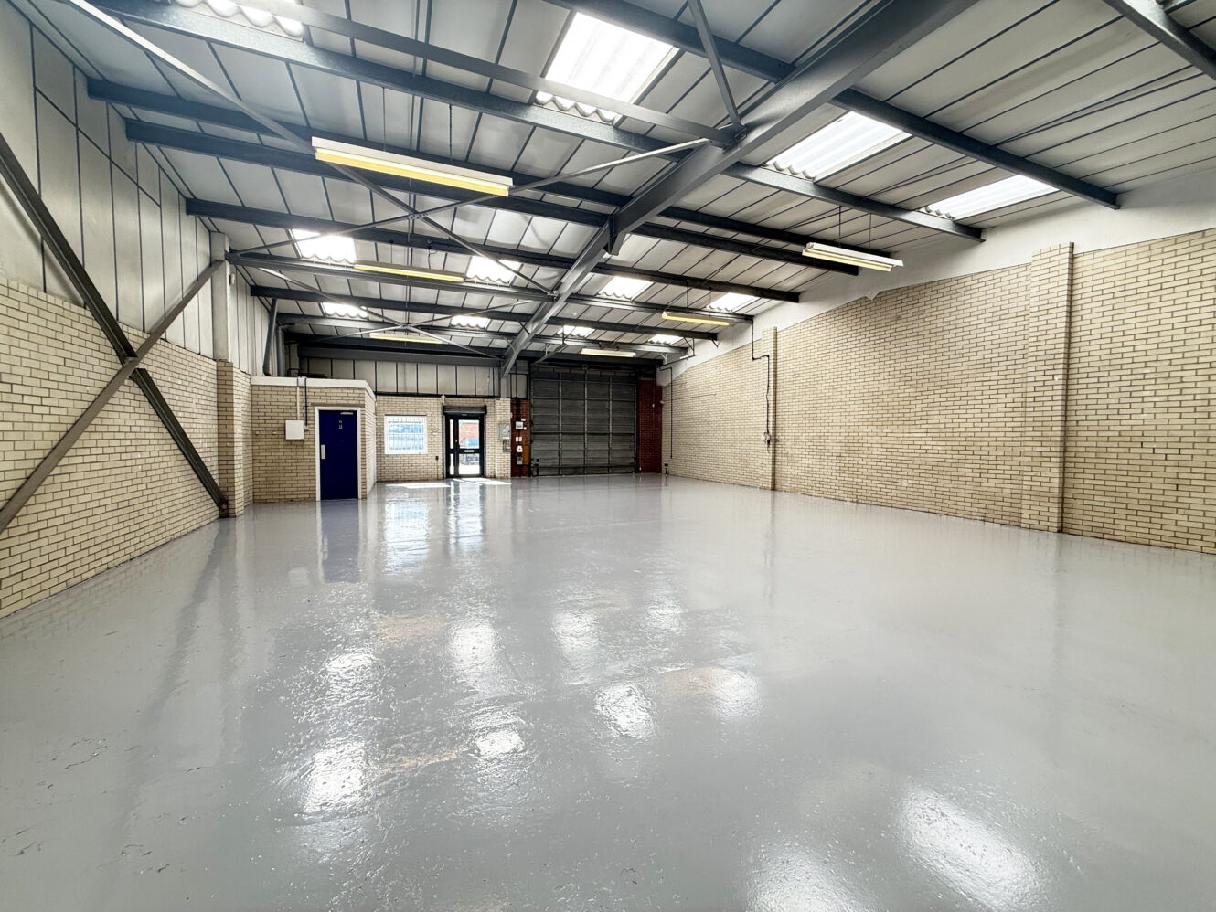 Empty industrial warehouse with brick walls, polished concrete floor, metal beams, fluorescent lights, blue door, and roller shutter.