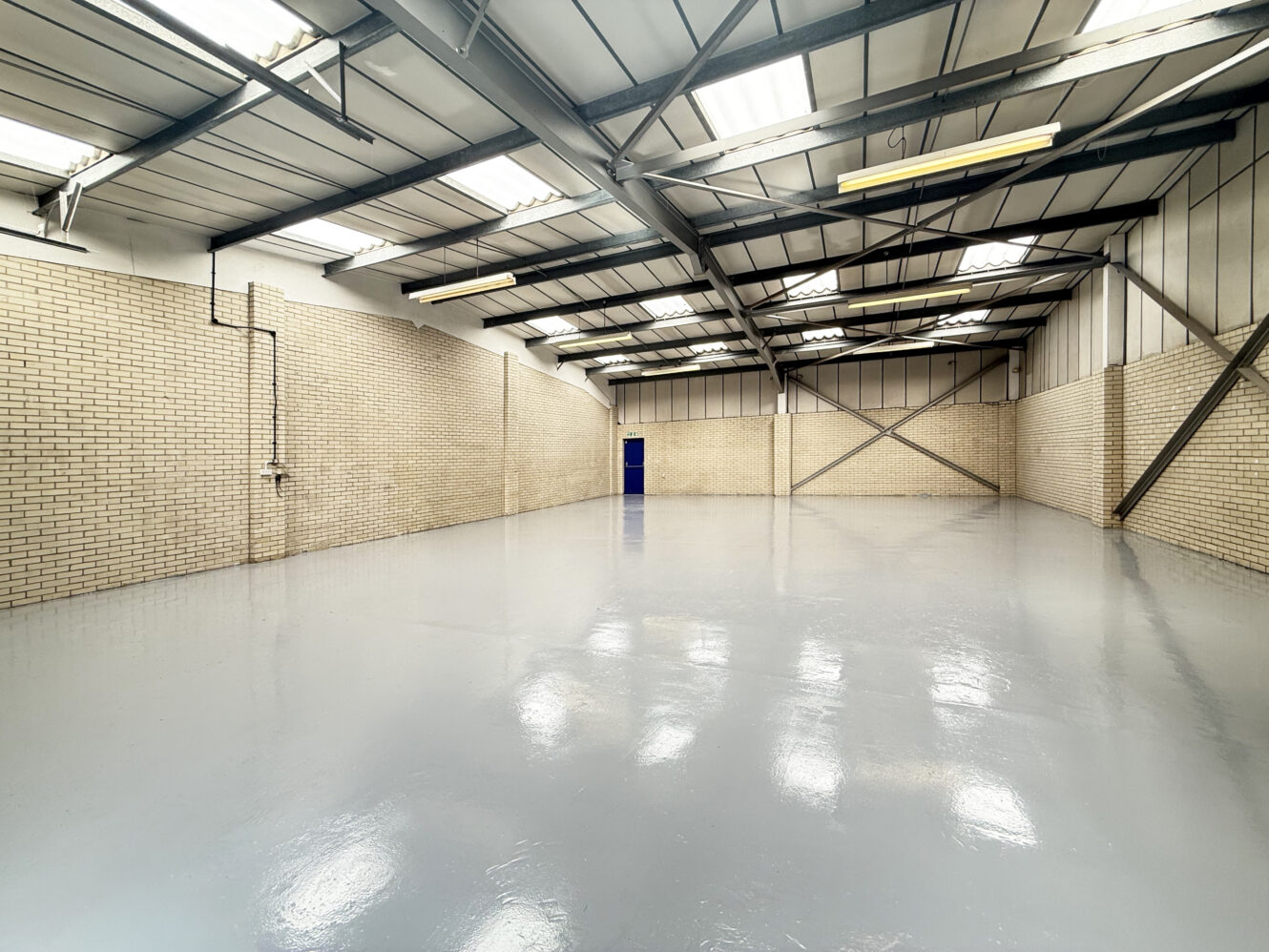 Empty industrial warehouse with brick walls, polished concrete floor, metal beams, fluorescent lights, and a blue door at the far end.