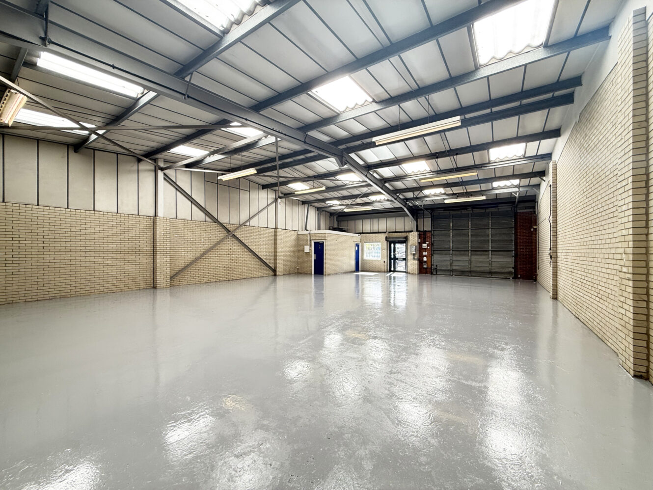 Empty industrial warehouse featuring a polished concrete floor, brick walls, steel beams, fluorescent lights, and a large roller shutter door.