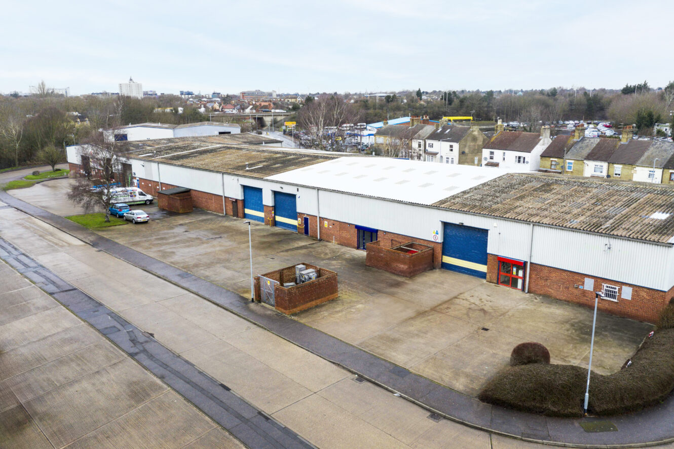 Aerial view of an industrial warehouse with blue doors, a large concrete yard, parked cars, trees, and surrounding buildings.