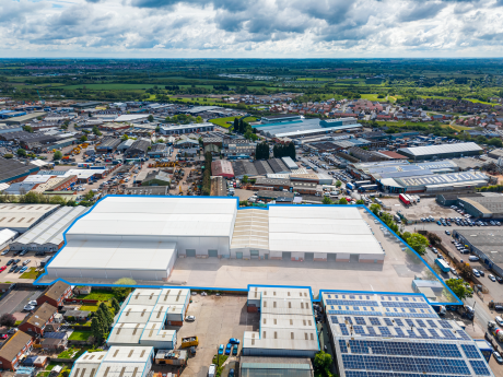 Aerial view of an industrial estate with large warehouses, smaller buildings, car parks, and residential areas under clouds.