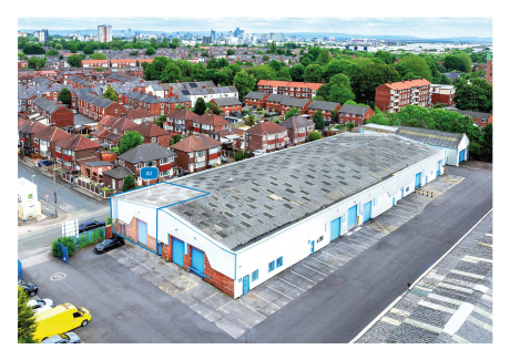 Aerial view of a large white warehouse with blue doors, surrounded by homes, trees, and several parked vehicles in an urban setting.