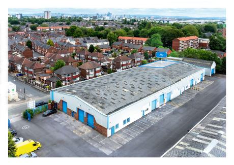 A large industrial building with blue doors stands beside brick houses in a residential area, with a city skyline in the distance.