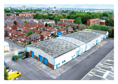 Aerial view of a large industrial warehouse with blue doors, surrounded by houses and trees in an urban setting.
