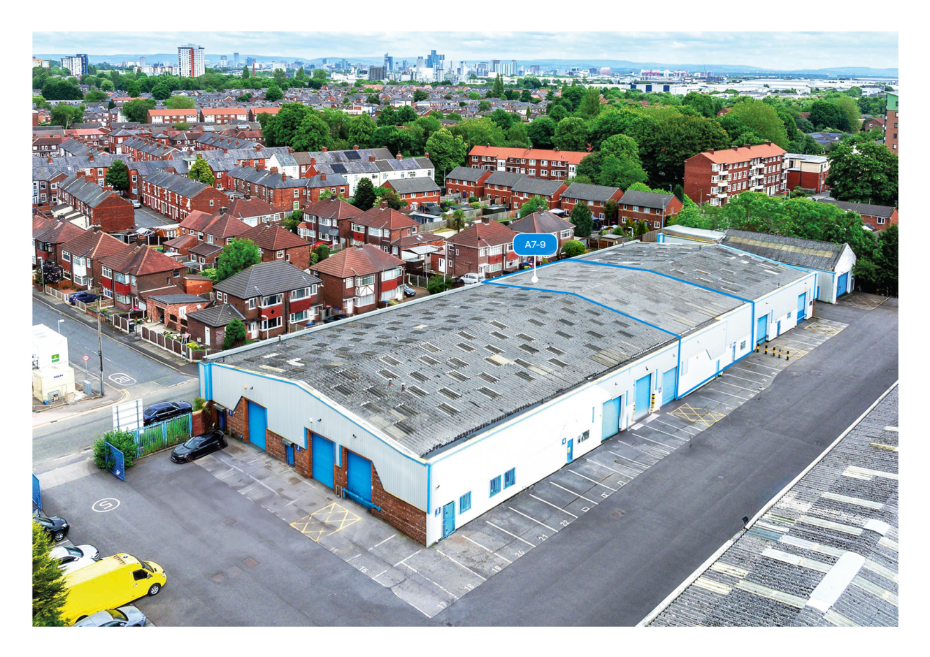 Aerial view of a large industrial warehouse with blue doors, next to homes and parked vehicles in an urban neighbourhood.