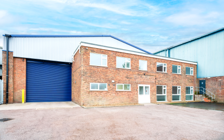 A two-storey brick industrial building with several windows, a white door, and a blue roller shutter beside a paved car park and cloudy sky.
