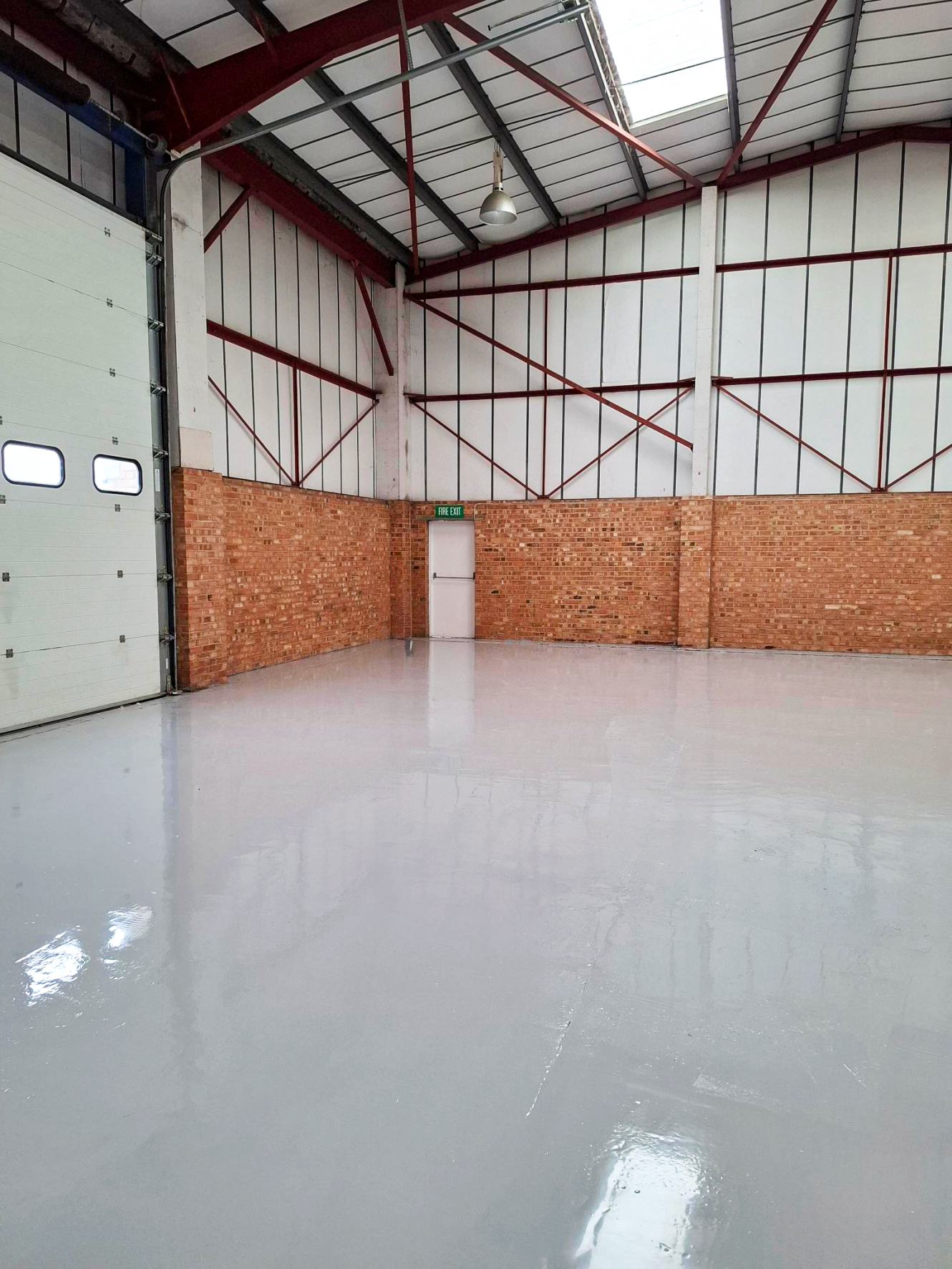 Interior of an empty warehouse with brick walls, high ceiling, shiny concrete floor, a white door, and a large industrial garage door.