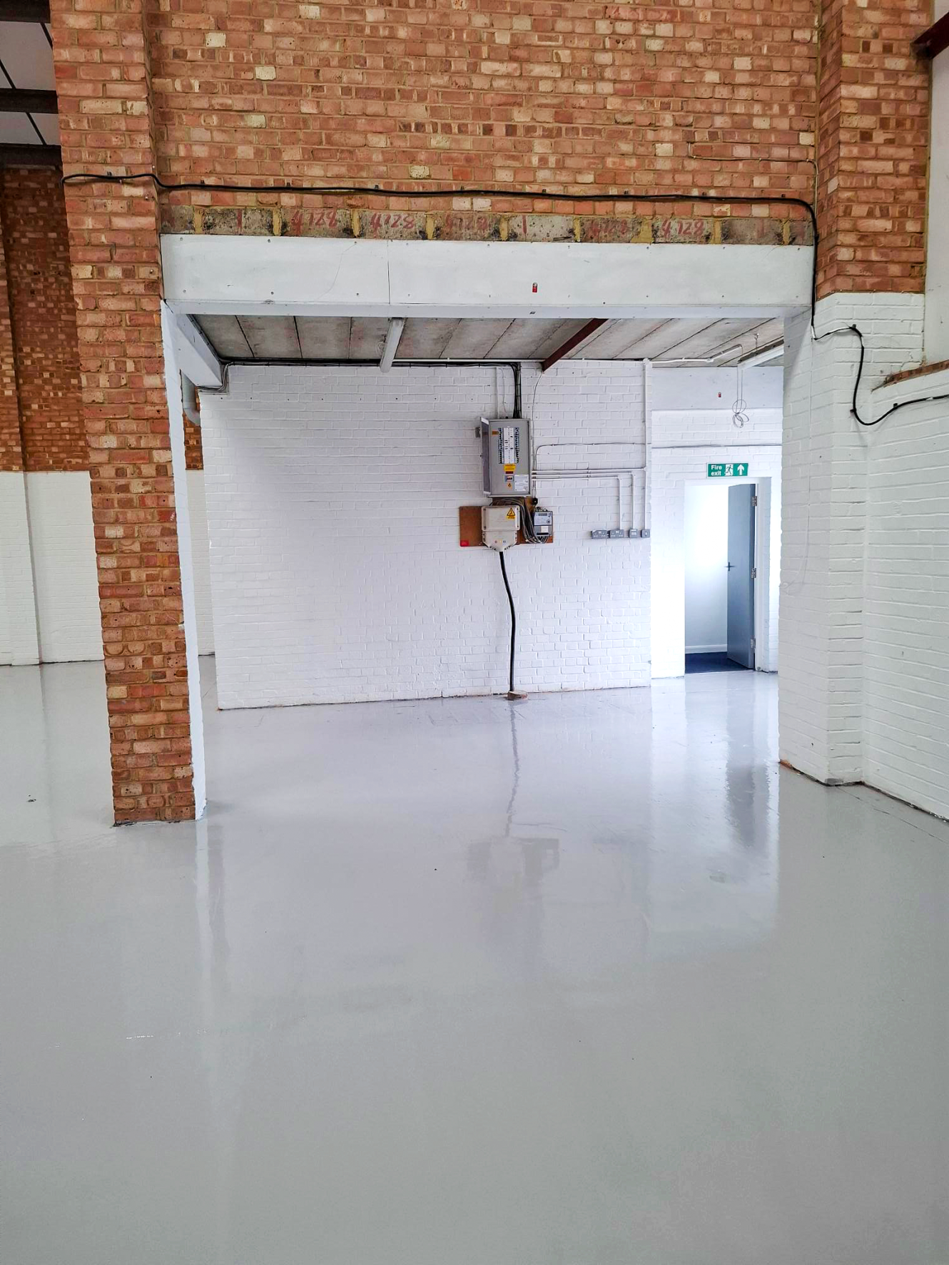 Empty industrial warehouse with brick walls, white floor, exposed ceiling, utility boxes, and open doorway to an exit sign.