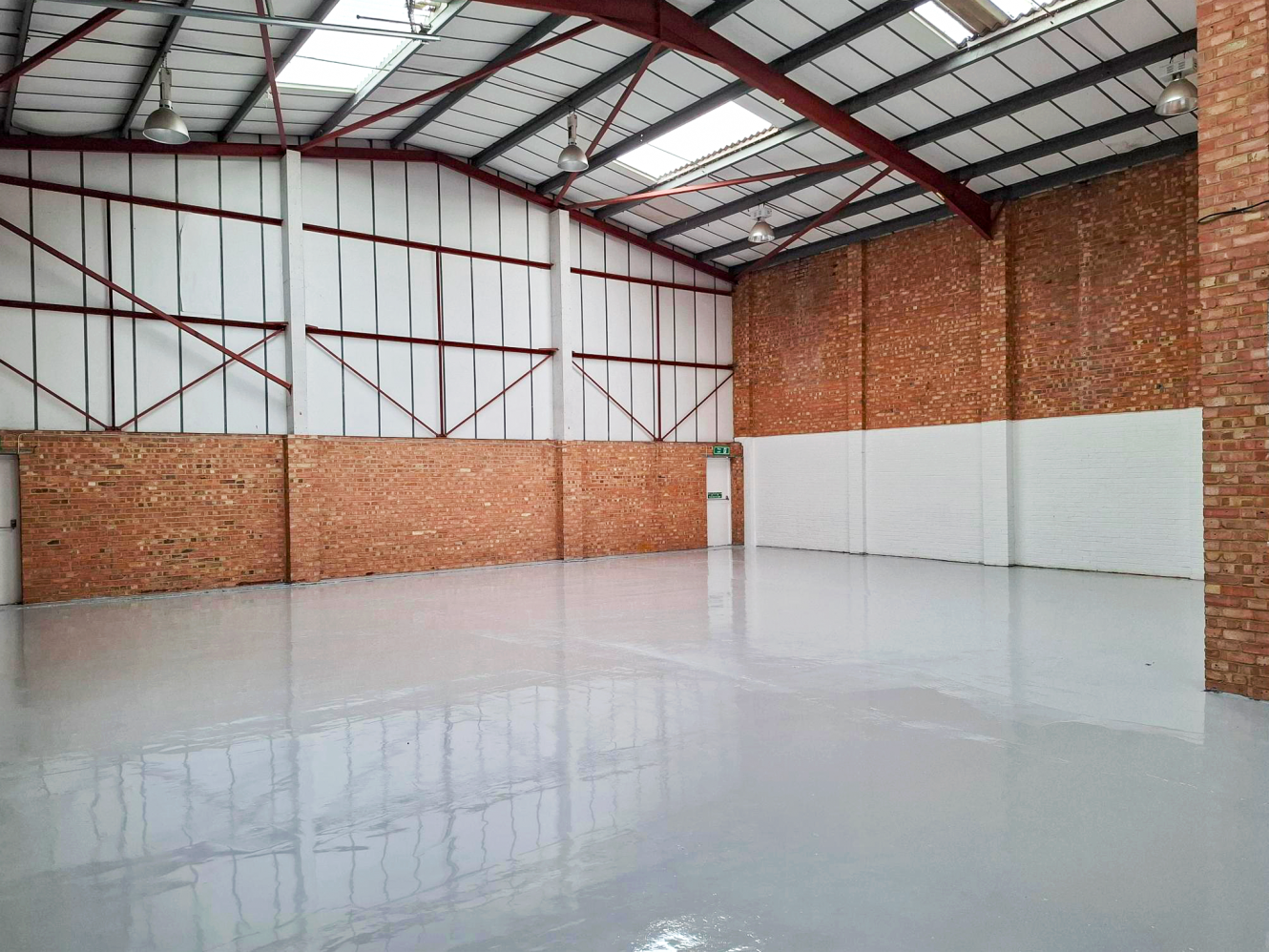 Empty industrial warehouse with brick walls, high metal ceiling, polished concrete floors, exposed beams, and a small door in the corner.