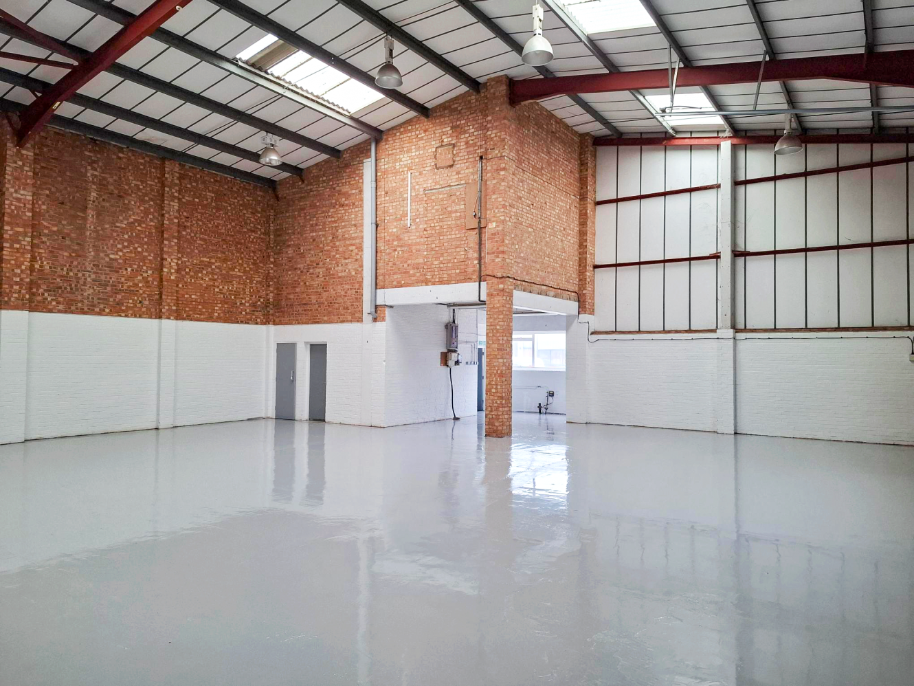 Interior of an empty warehouse with red steel beams, brick and white-painted walls, glossy floor, and skylights overhead.