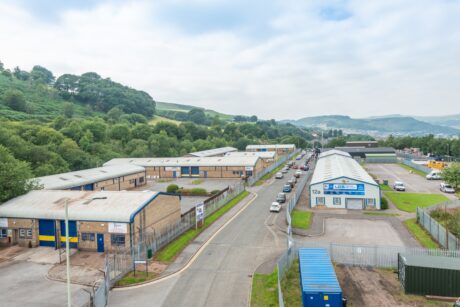 Aerial view of an industrial estate with warehouses, a fenced road, parked cars, and green hills under a partly cloudy sky.