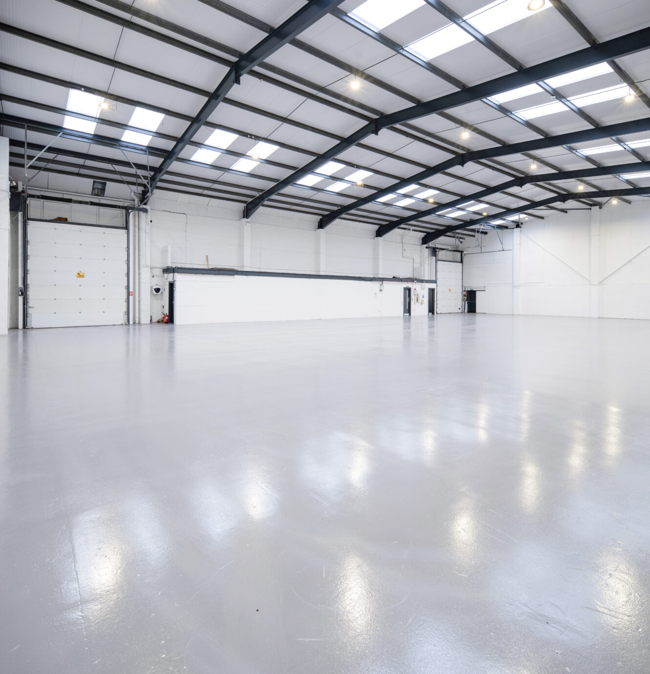 Spacious warehouse with white walls, polished concrete floor, high ceiling, skylights, and several garage doors.