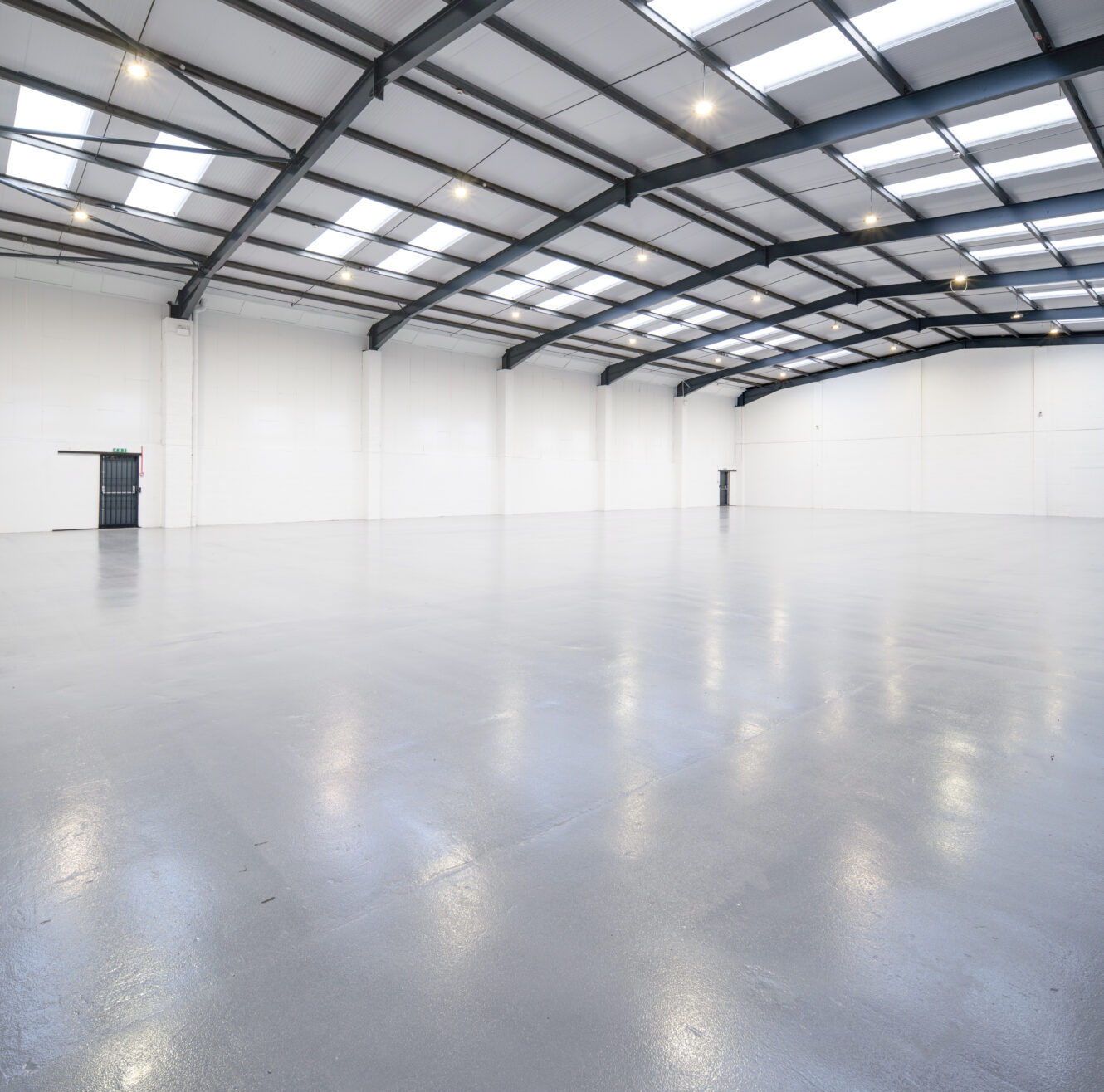 Large, empty warehouse with polished concrete floor, white walls, and a high ceiling featuring metal beams and rooflights.