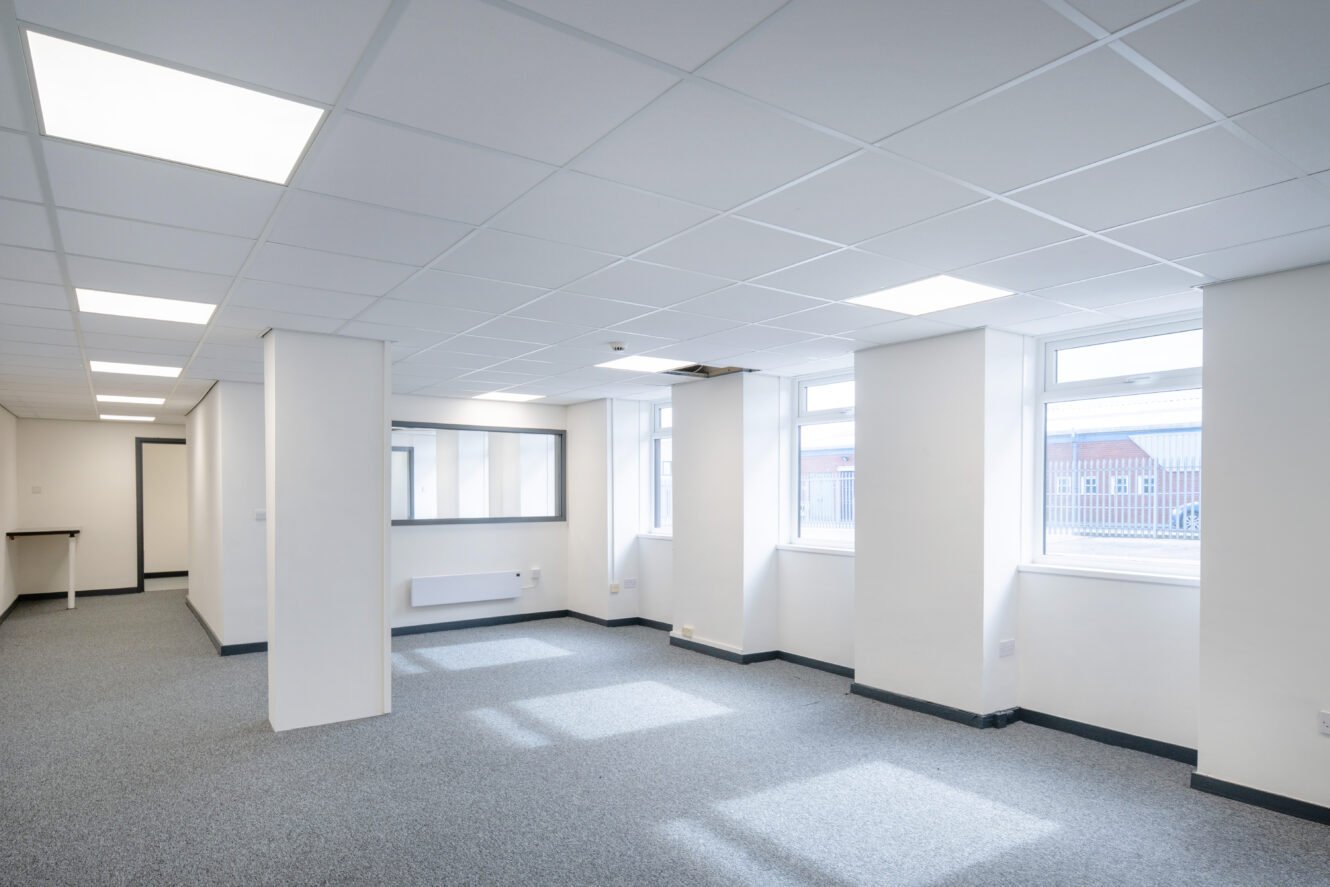 Empty, well-lit office space with white walls, grey carpet, multiple windows, ceiling lights, columns, and a glass partition.