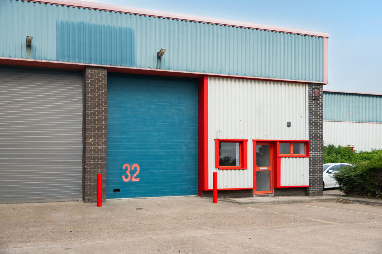 Industrial warehouse unit with blue roller shutter labelled 32, smaller grey door, red trim, office window, and car partly visible on right.