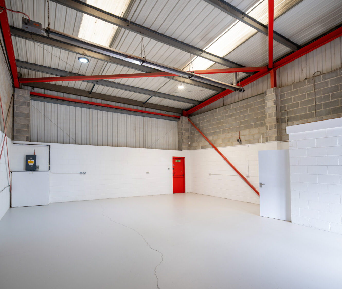 Empty industrial warehouse with white floors and walls, exposed beams, red door, open white door, ceiling lights, and electrical panels.