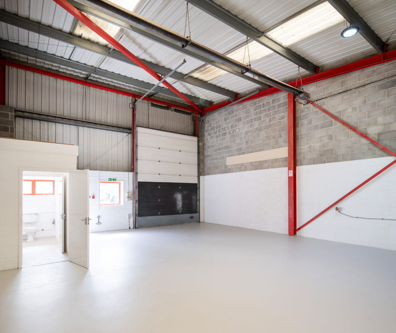 Interior of an empty industrial warehouse with painted walls, exposed beams, a roller shutter door, and a small room with an open door.