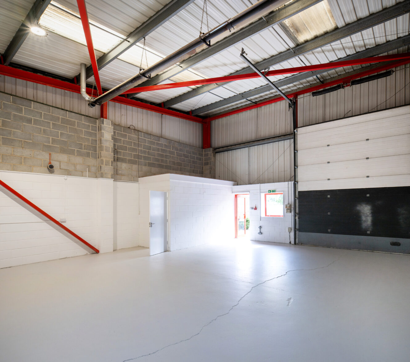 Empty warehouse with painted concrete floor, exposed brickwork, steel beams, white office, shutter door, and natural light.