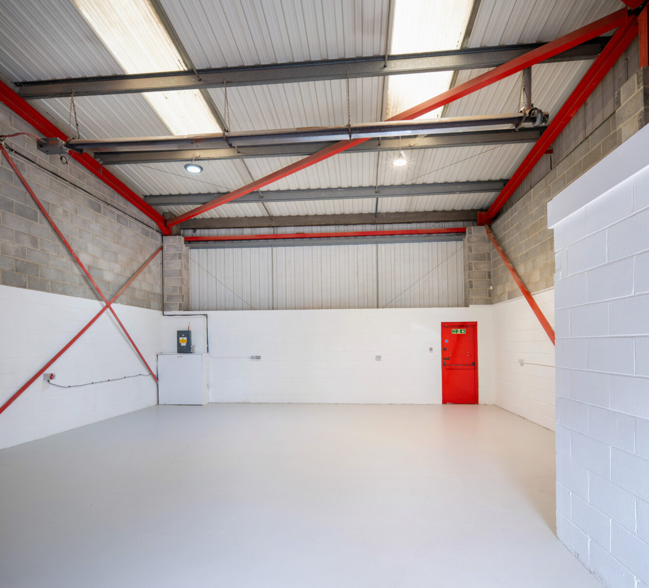 Empty industrial warehouse space with white walls, exposed brickwork, red steel beams, high ceiling, and a red door on the right wall.