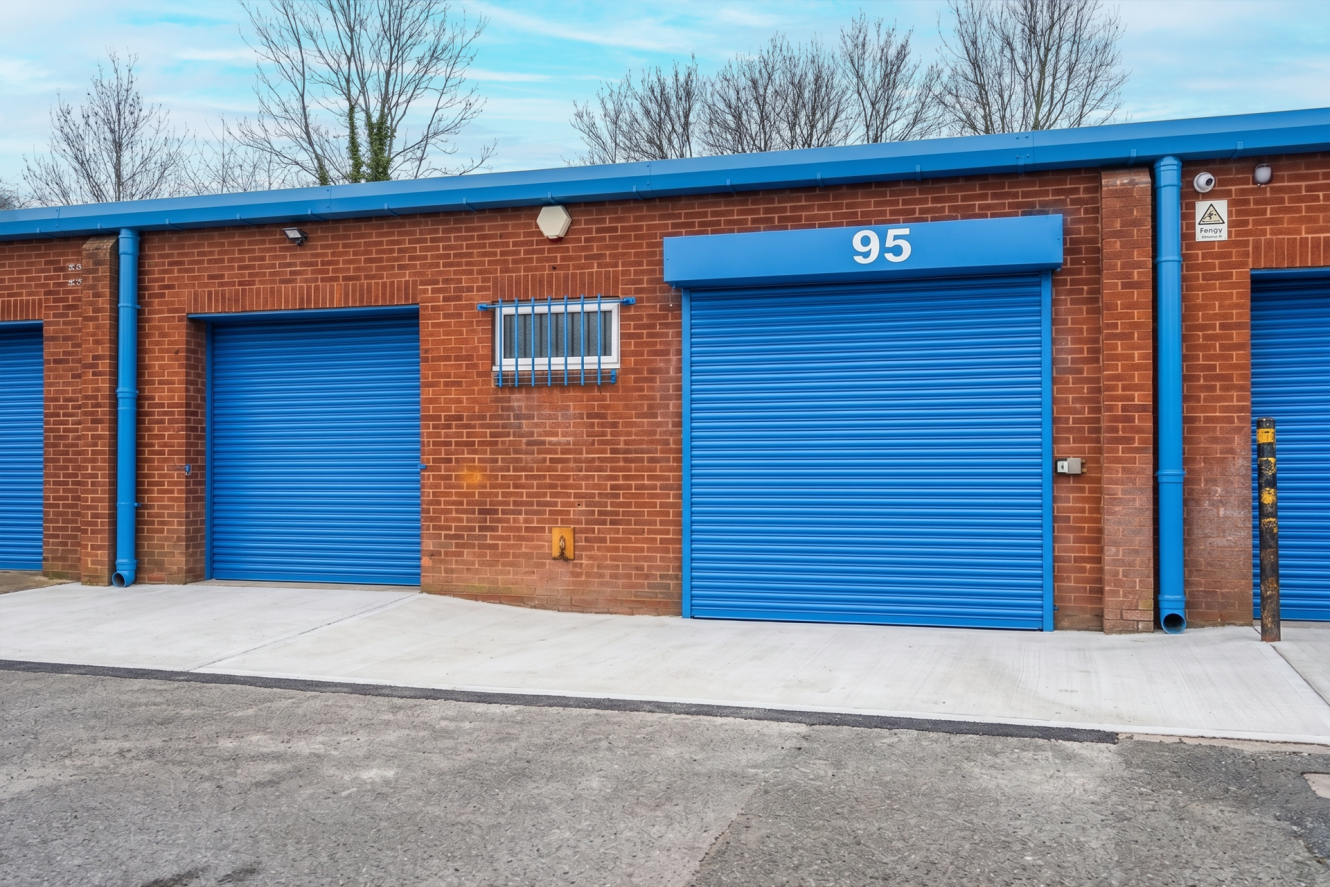 A row of red brick storage units with blue roller doors; unit 95 is centred. Leafless trees stand in the background.