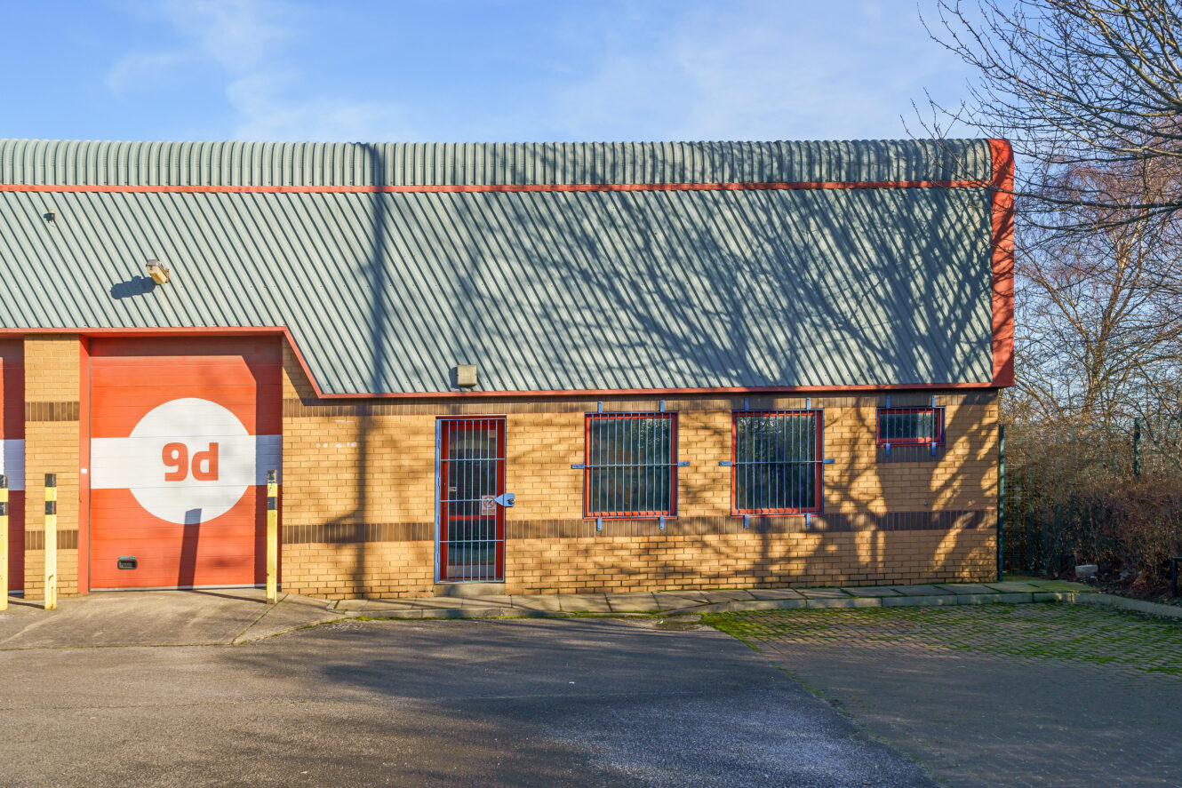 Industrial building with corrugated metal roof, brick walls, and a red garage door labelled 9d; tree branch shadows on facade.