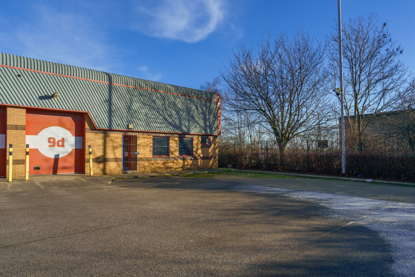Industrial building with a red garage door labelled 9d, large windows, and an empty car park under a clear sky.
