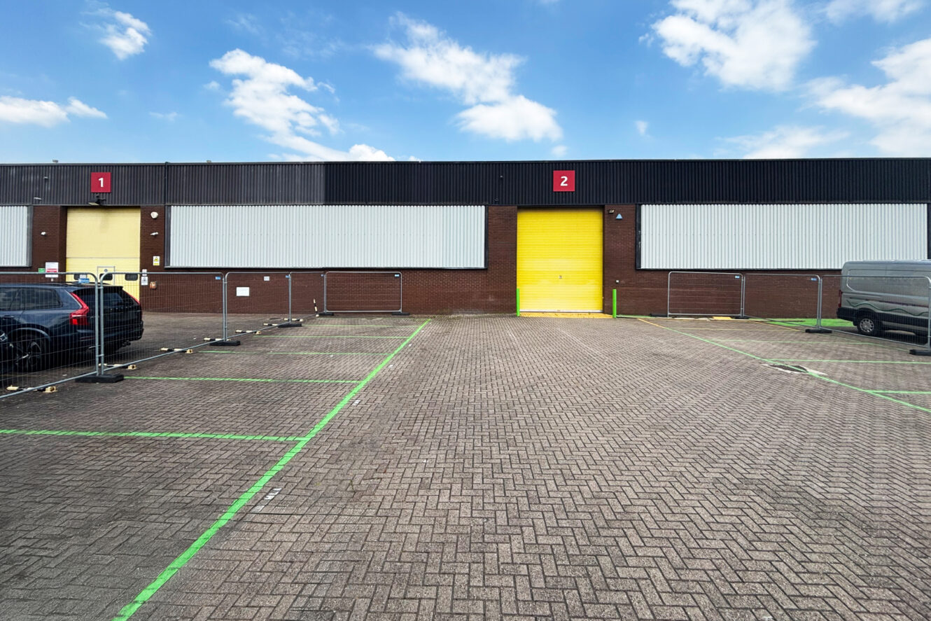 A warehouse with a yellow door labelled 2 next to a car park with green-marked spaces and several vehicles, under a cloudy sky.