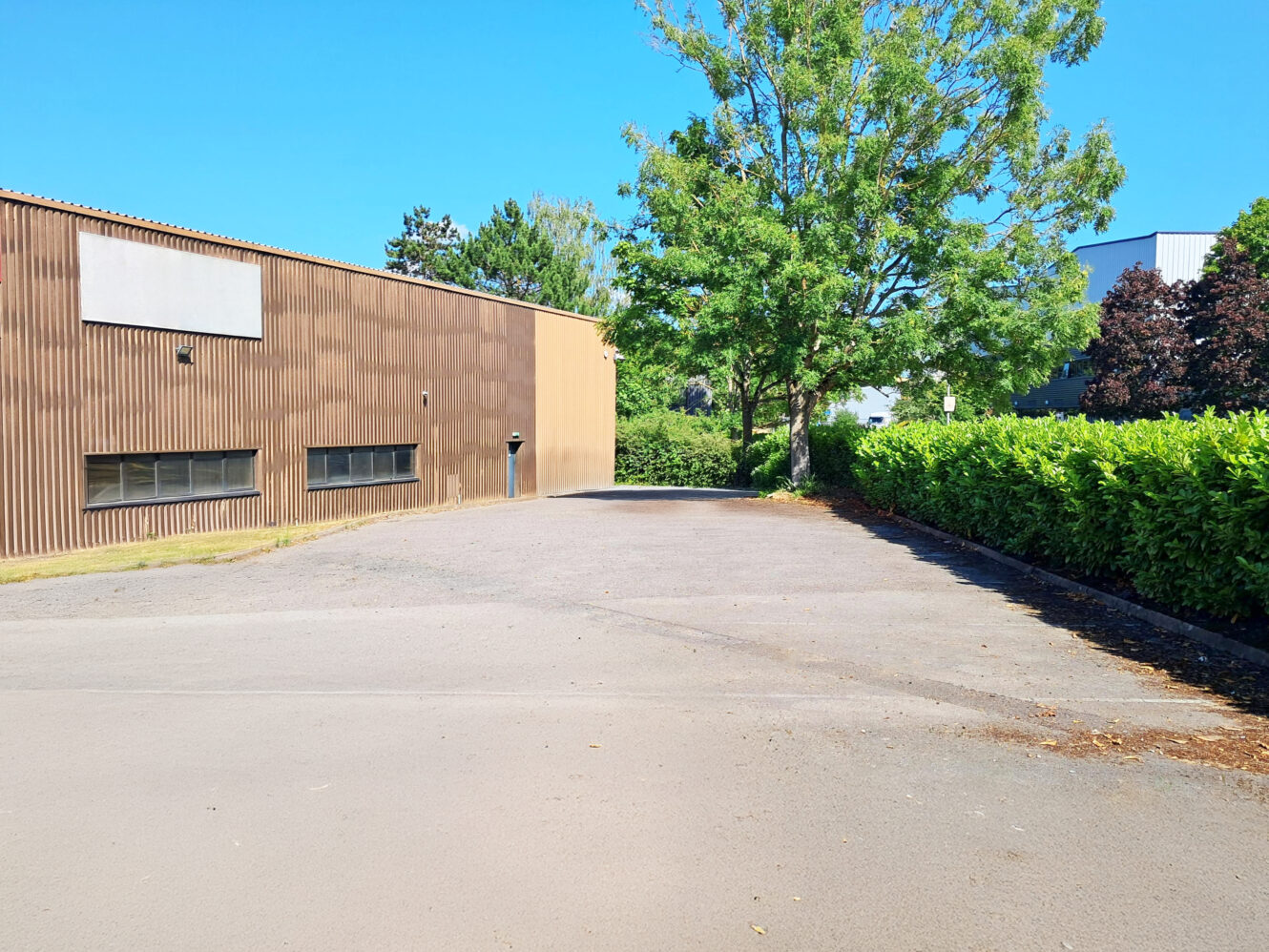 Empty paved space next to a brown industrial building, lined with green hedges and trees, beneath a clear blue sky.