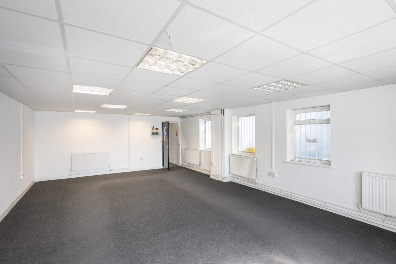 Empty office room with white walls, ceiling lights, barred windows, radiators, and dark carpet flooring.