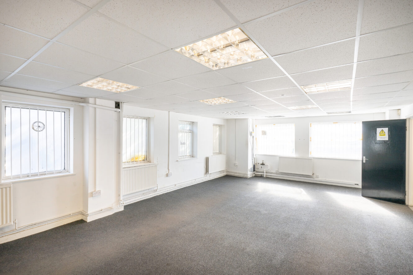 A brightly lit office room with white walls, grey carpet, several windows, and fluorescent ceiling lights.