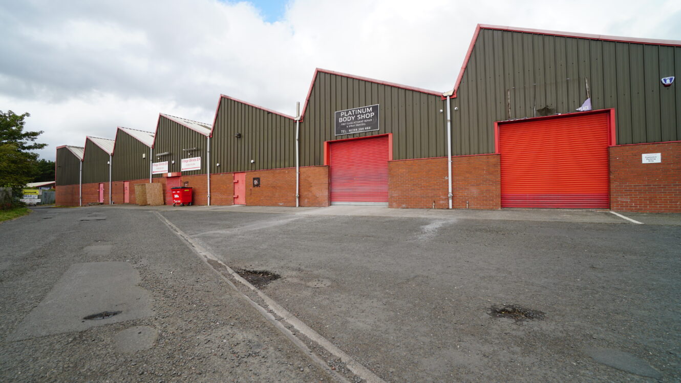 A row of industrial warehouse units with green cladding, red brick bases, and red roller shutters along a tarmacked road under a cloudy sky.