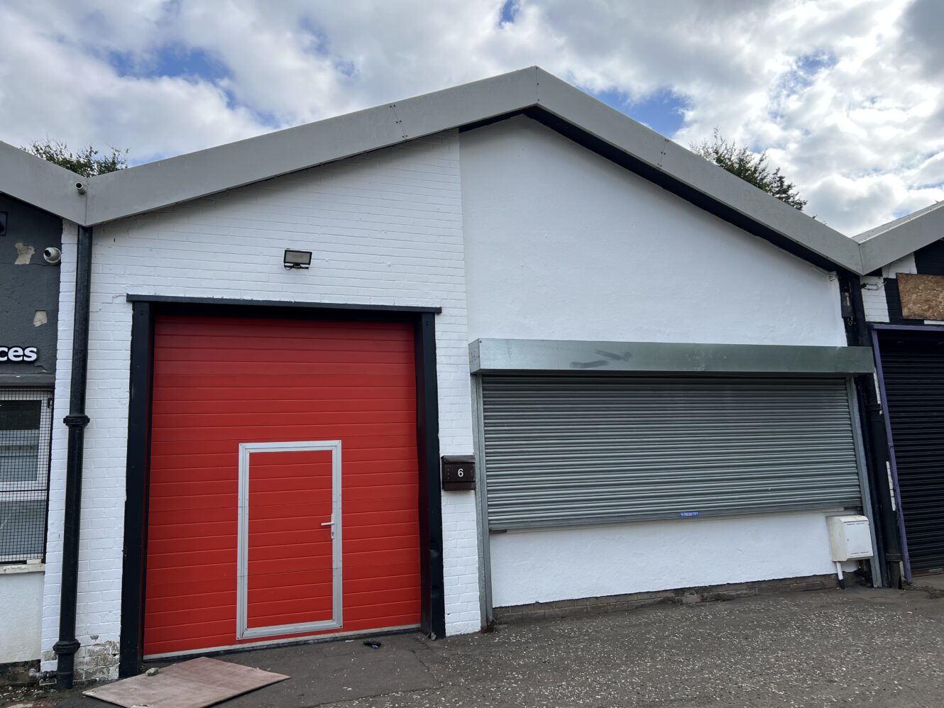 A white industrial building with a large red door on the left, a closed silver roller shutter on the right, and postbox 6 mounted between them.