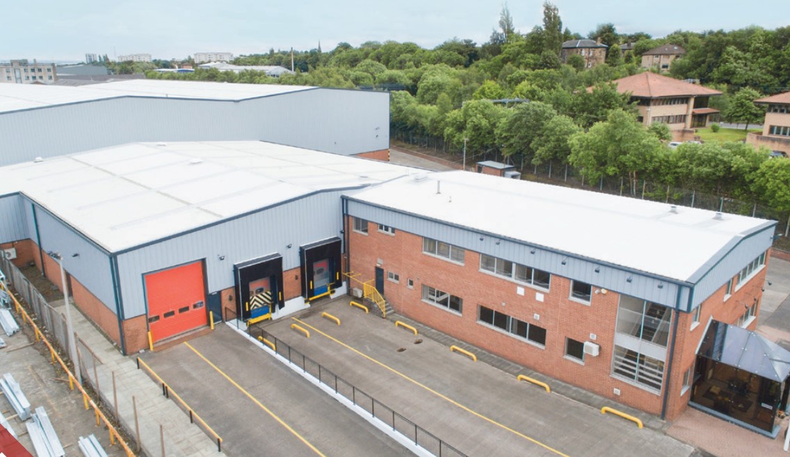 A large industrial warehouse with two loading bays and a brick office section, surrounded by trees and nearby buildings.