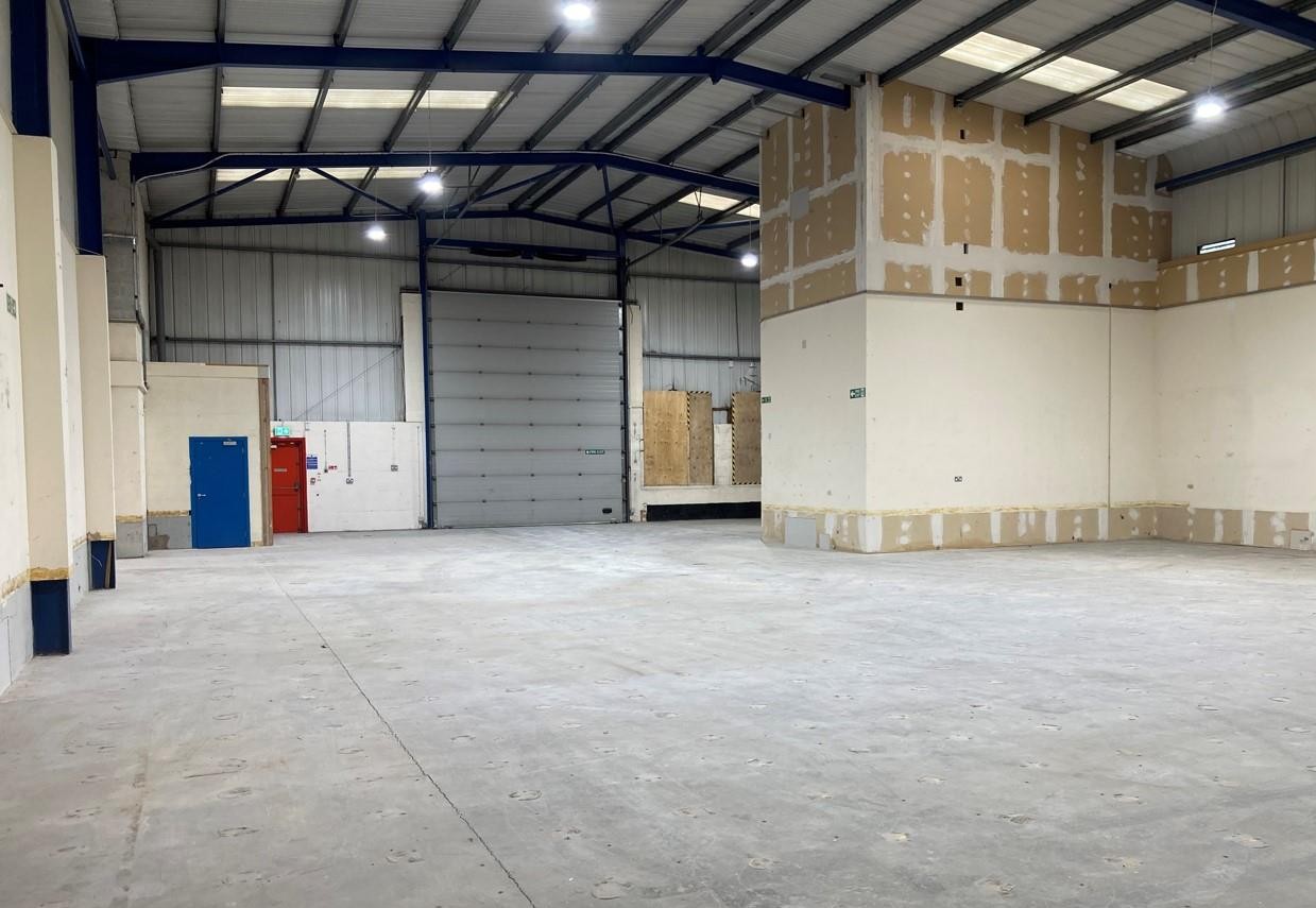 Empty warehouse interior with high ceilings, exposed beams, large roller shutter, unfinished walls, and patches of plasterboard on concrete floor.