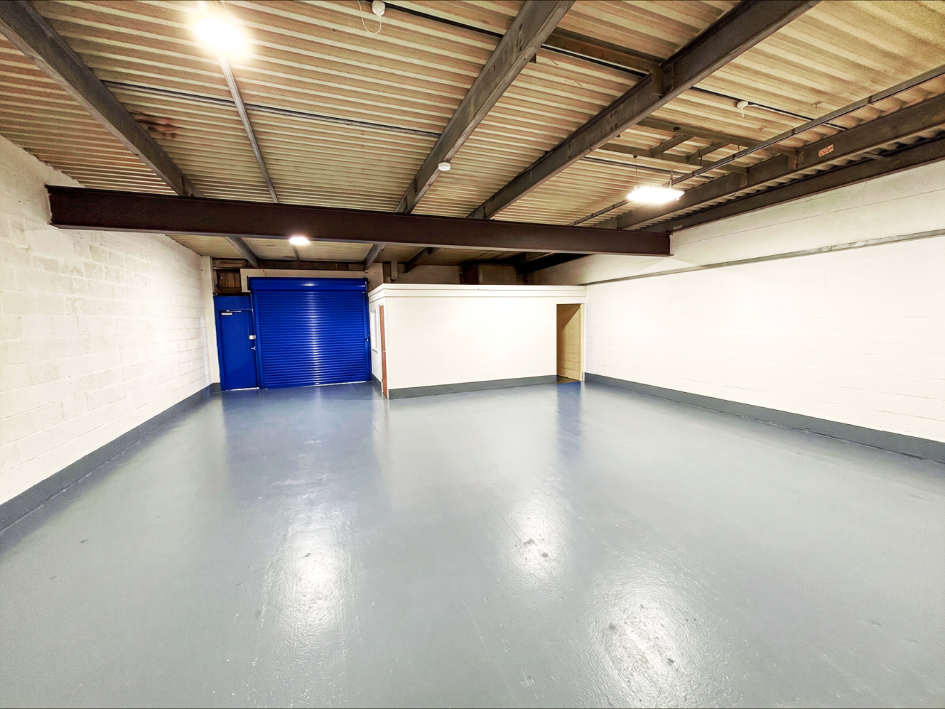 Empty industrial unit with painted concrete floor, white brick walls, exposed beams, blue roller shutter, fluorescent lights, and enclosed room.
