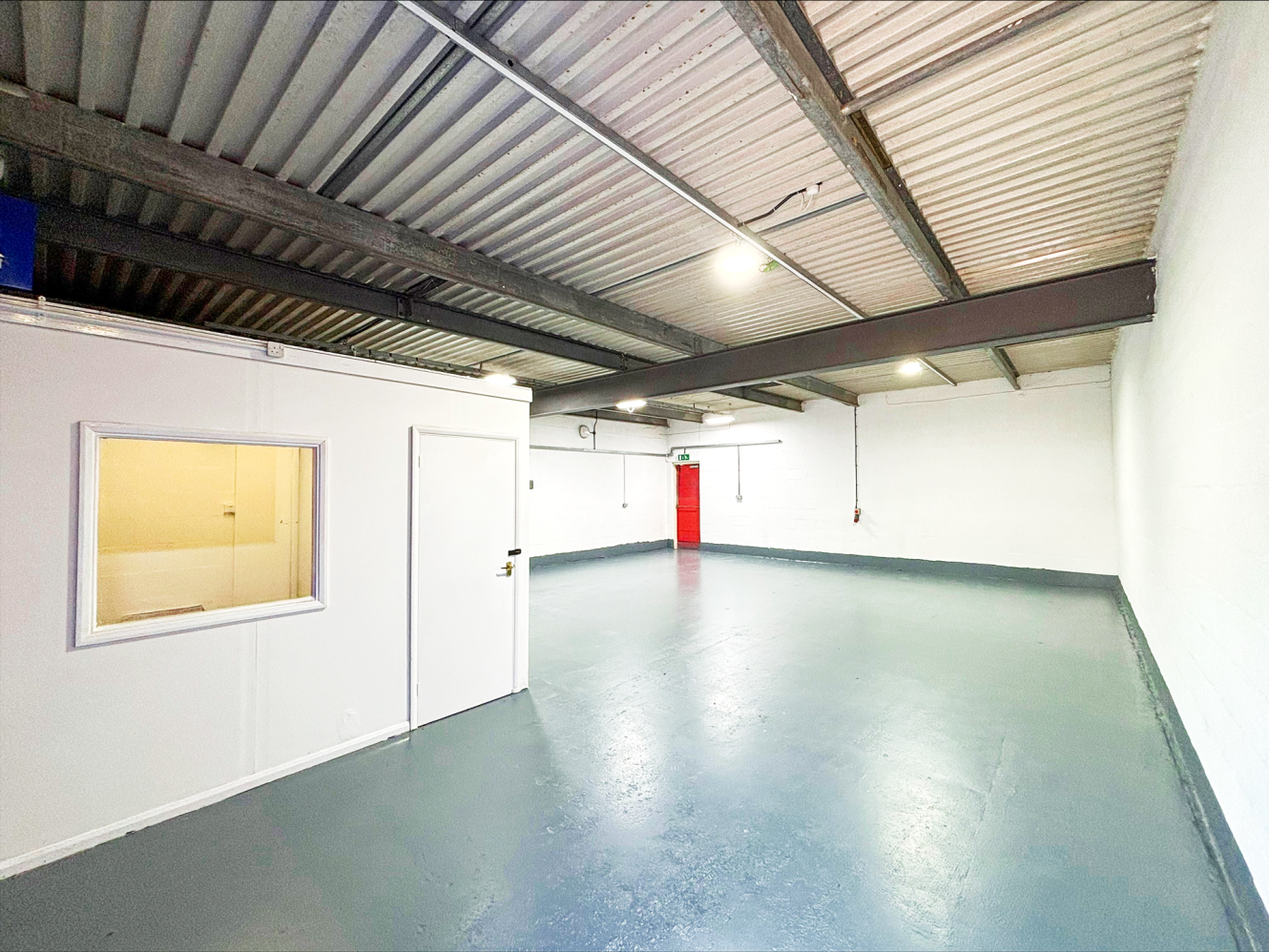 Empty industrial room with grey floors, white walls, exposed metal ceiling, a small white office, and a red exit door at the back.