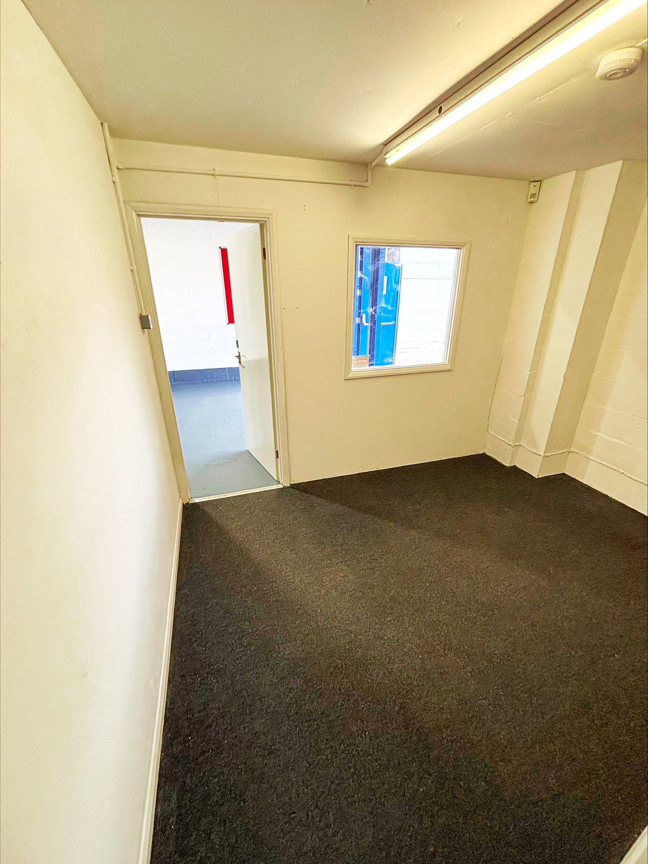 Small, empty room with white walls, dark carpeted floor, a window, and an open door to another brightly lit area.