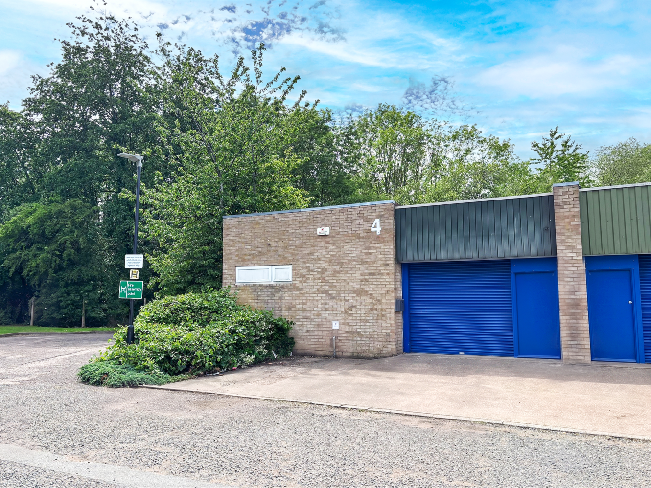 A single-storey brick building with blue doors numbered “4”, beside trees and a signpost on a tarmacked area under a partly cloudy sky.